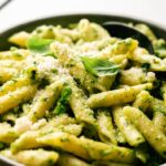 A close-up of Pesto Pasta, with penne tossed in green pesto sauce, garnished with grated parmesan cheese and fresh basil leaves, served in a bowl with a black spoon.
