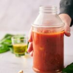 A hand holds a glass jar filled with pizza sauce; a small glass of olive oil and fresh basil leaves are in the background.