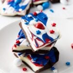 A stack of Red White and Blue Chocolate Bark with swirled white, blue, and red topping, decorated with star-shaped sprinkles and candy pieces, sits on a white plate.