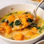 A bowl of creamy Sweet Potato Soup with chunks of butternut squash, spinach, and onions, with a spoon lifting a portion.