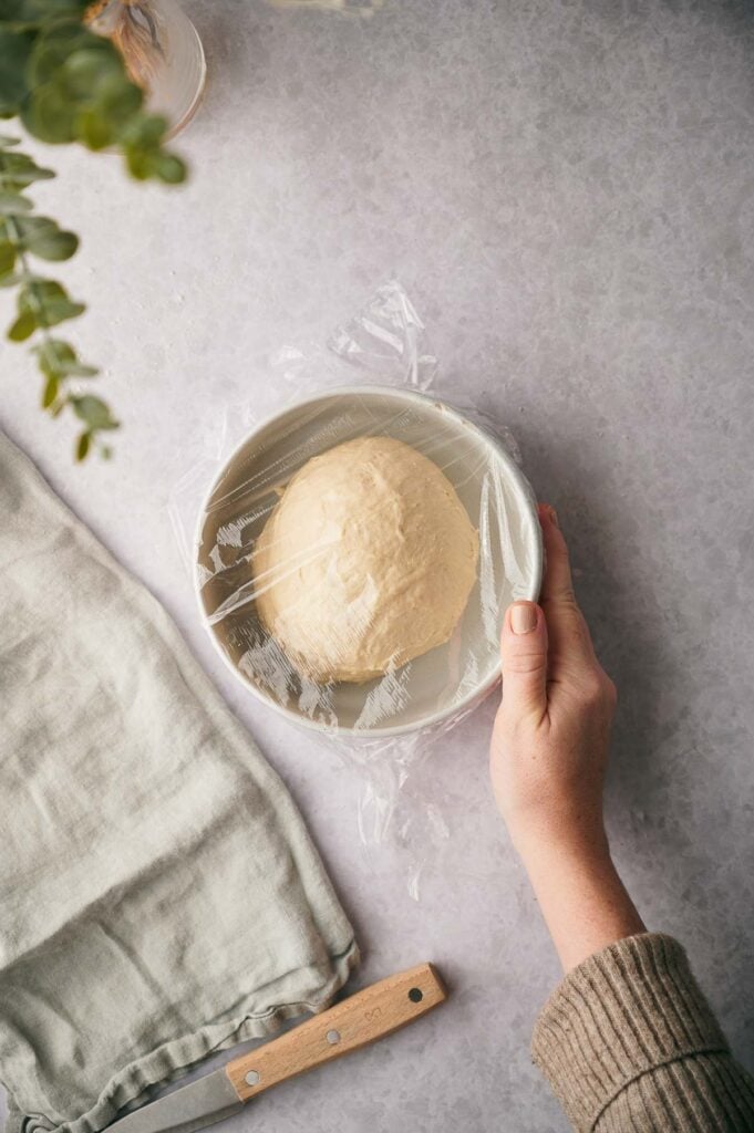 A hand covers a bowl of dough with plastic wrap on a gray countertop, preparing it for a flatbread recipe; a folded cloth and a knife rest nearby.