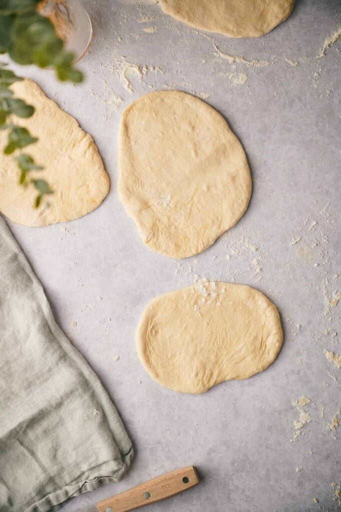 Three pieces of rolled-out dough for a flatbread recipe rest on a floured surface, with a cloth and a plant partially visible on the left side.