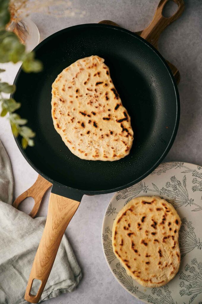 A flatbread sizzles in a nonstick frying pan with a wooden handle, while another freshly cooked flatbread rests on a patterned plate beside the pan.
