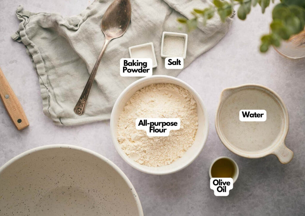 A bowl of flour, baking powder, salt, water, and olive oil are arranged on a countertop with a spoon and cloth&mdash;perfect for making homemade flatbread.