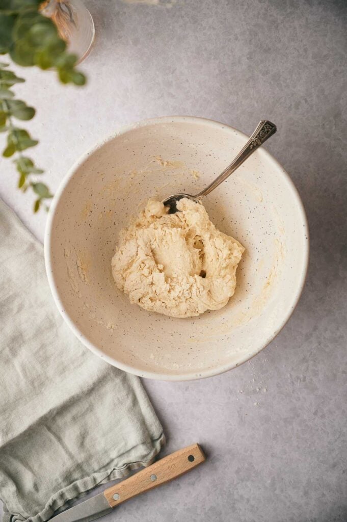 A white bowl with a spoon holds dough for a flatbread recipe on a light surface. A folded cloth and a knife are nearby, while part of a potted plant appears in the corner.