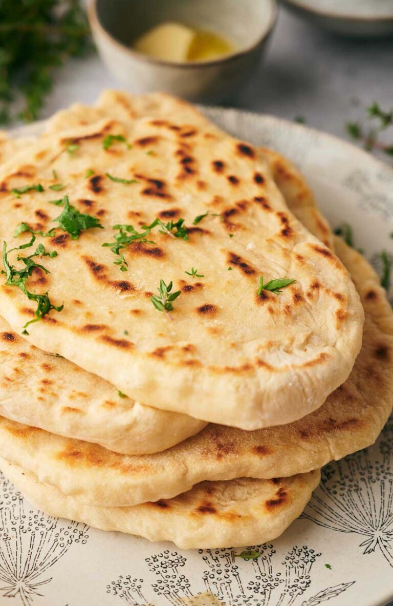 A stack of freshly cooked flatbread naan garnished with chopped herbs, placed on a patterned plate with a small bowl of butter in the background.