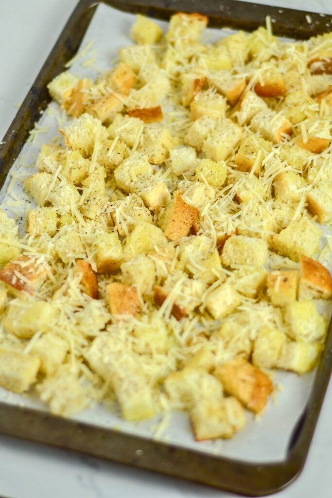A baking sheet lined with parchment paper, topped with cubed bread and grated cheese, is ready to bake into crispy Cheese Croutons.