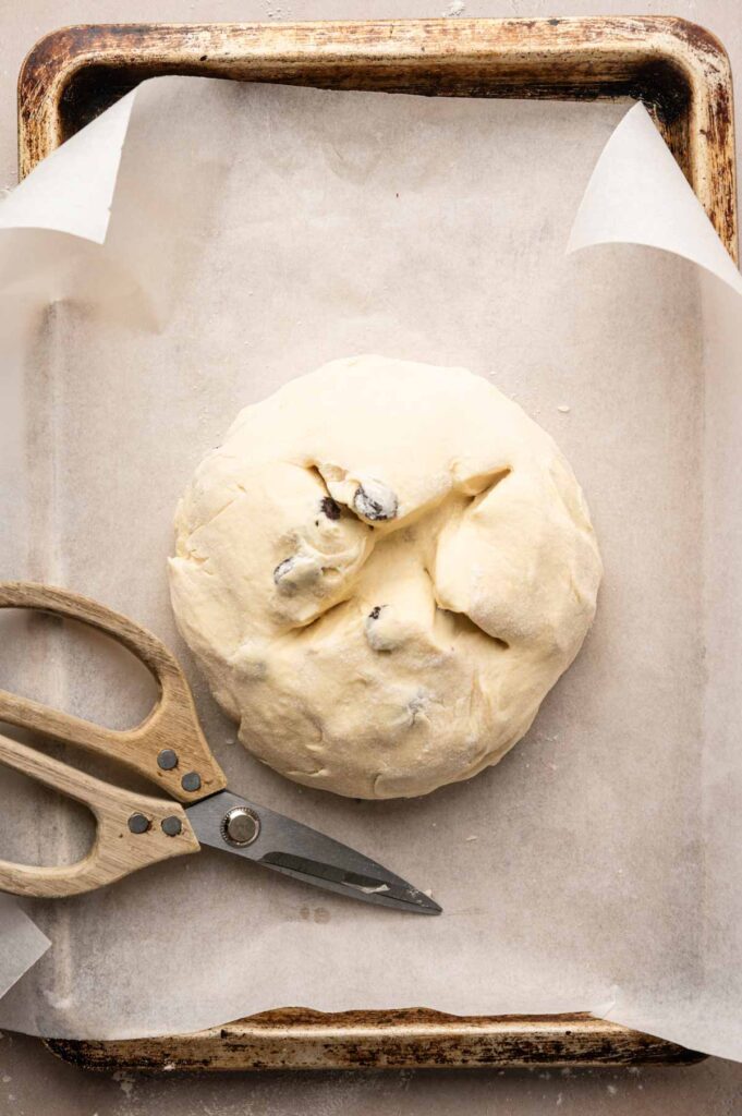 Round ball of Irish Soda Bread dough with raisins on a parchment-lined baking sheet, next to a pair of wooden-handled kitchen scissors.