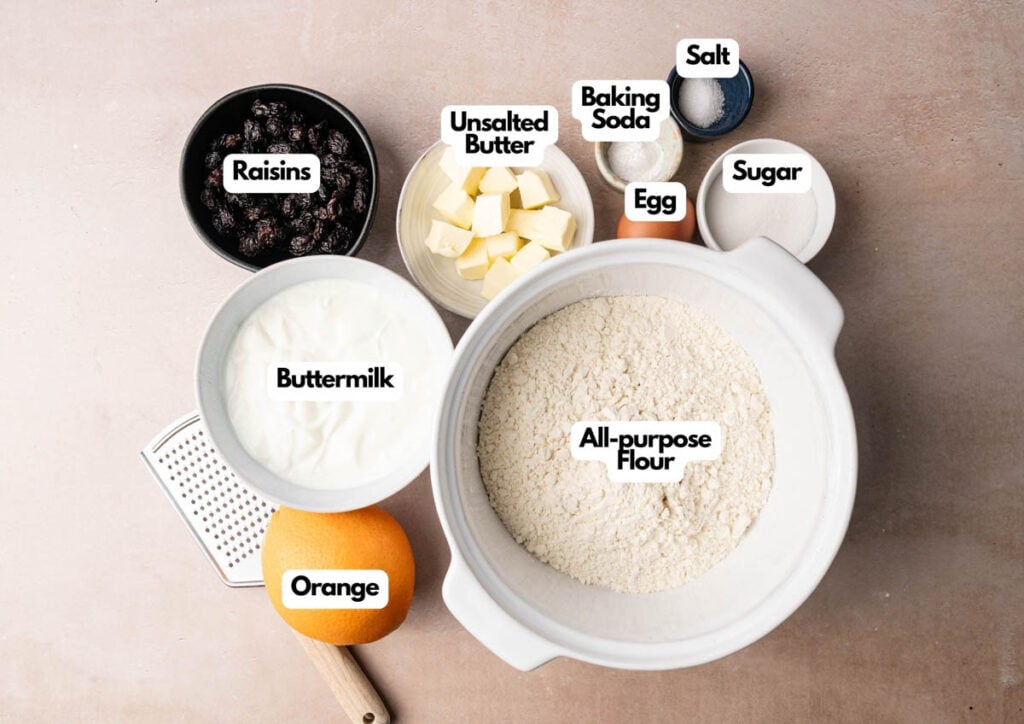 Labeled ingredients for baking Irish Soda Bread, including all-purpose flour, buttermilk, orange, raisins, unsalted butter, egg, sugar, salt, and baking soda arranged on a countertop.