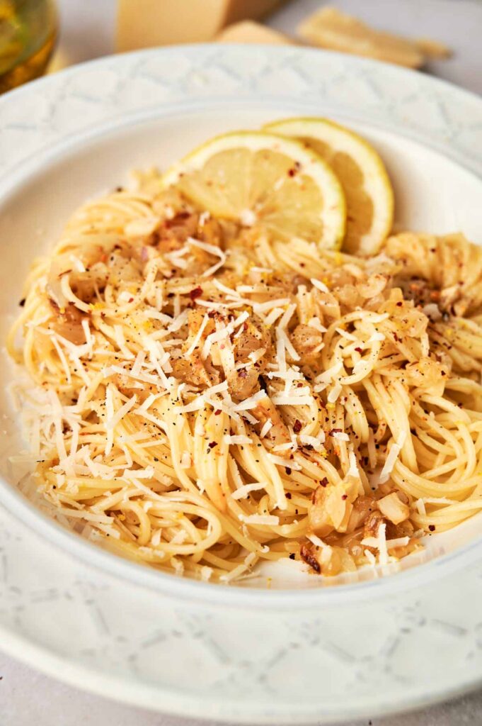 A plate of Lemon Pasta topped with grated cheese, saut&eacute;ed onions, and black pepper, garnished with fresh lemon slices.