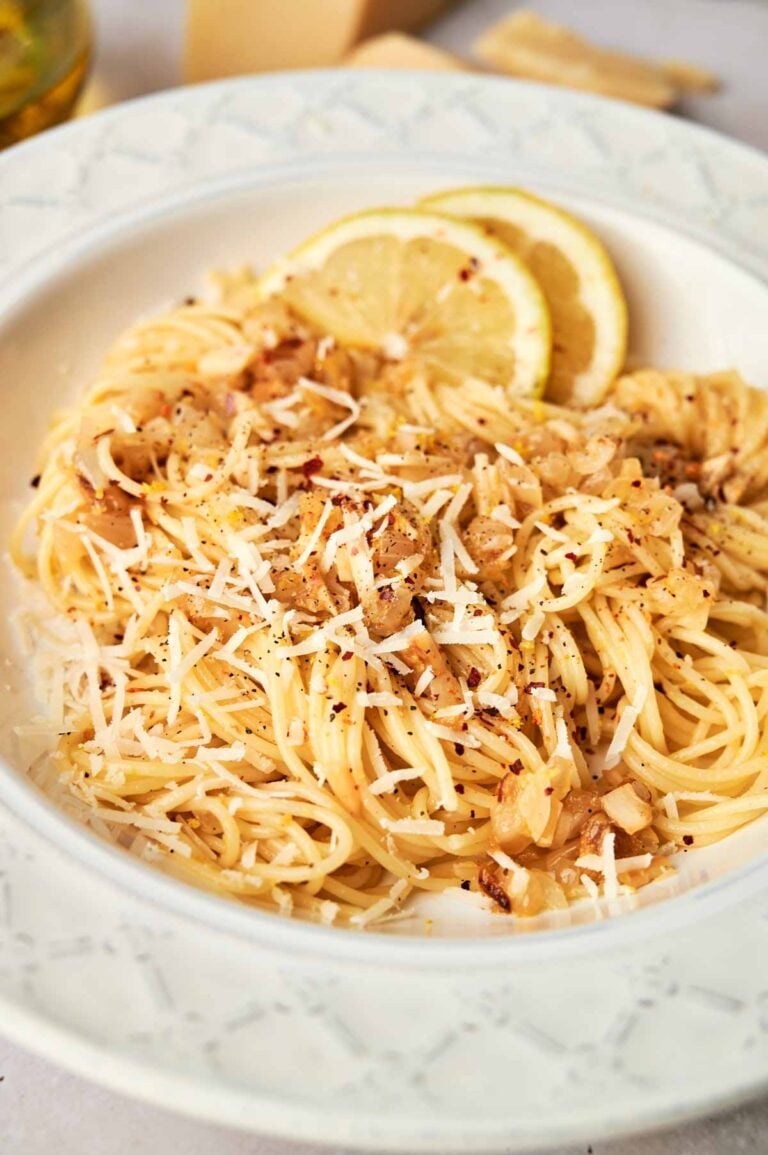 A plate of Lemon Pasta topped with grated cheese, sautéed onions, and black pepper, garnished with fresh lemon slices.