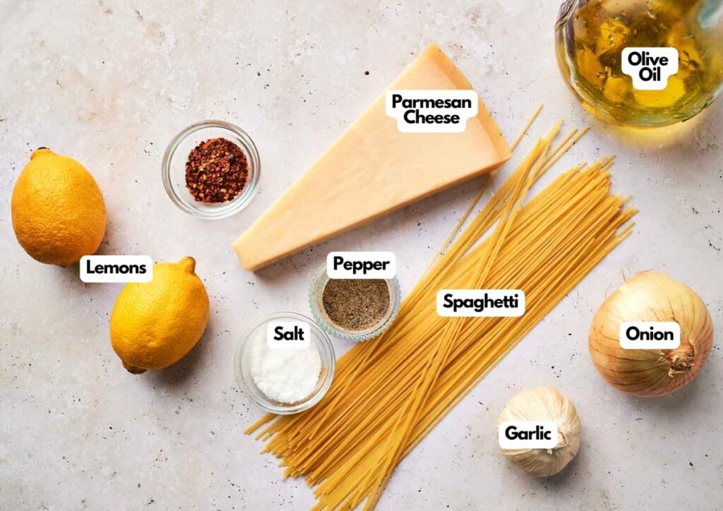A top-down view of Lemon Pasta ingredients: lemons, Parmesan cheese, olive oil, spaghetti, onion, garlic, salt, pepper, and red pepper flakes on a light surface.