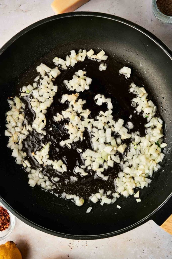 Chopped onions are being sautéed in oil in a black frying pan on a light-colored countertop, creating an aromatic base perfect for flavorful dishes like lemon pasta.