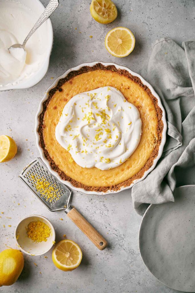 A Lemon Pie tart with whipped cream and lemon zest sits on a gray surface, surrounded by halved lemons, a grater with zest, a bowl of whipped cream, and a gray napkin.