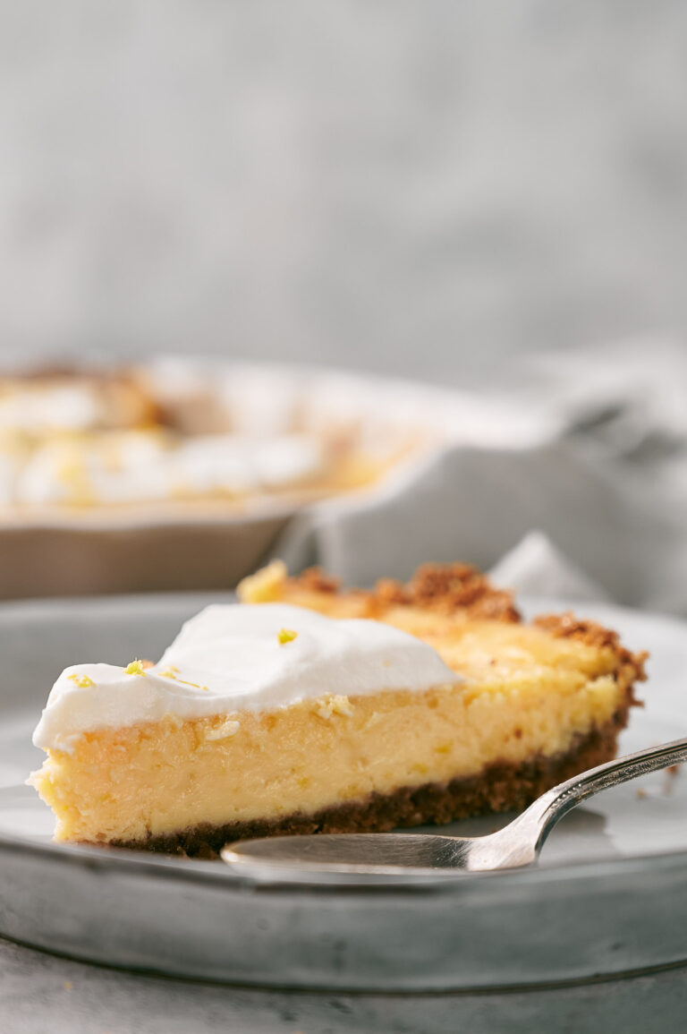 A slice of creamy lemon pie with a golden crust and a layer of whipped topping sits on a plate with a fork.
