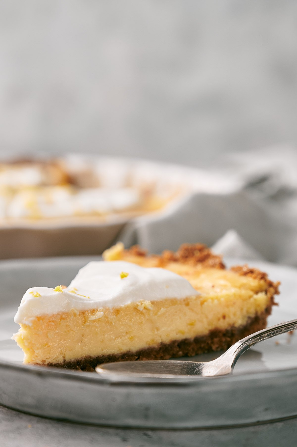 A slice of creamy lemon pie with a golden crust and a layer of whipped topping sits on a plate with a fork.