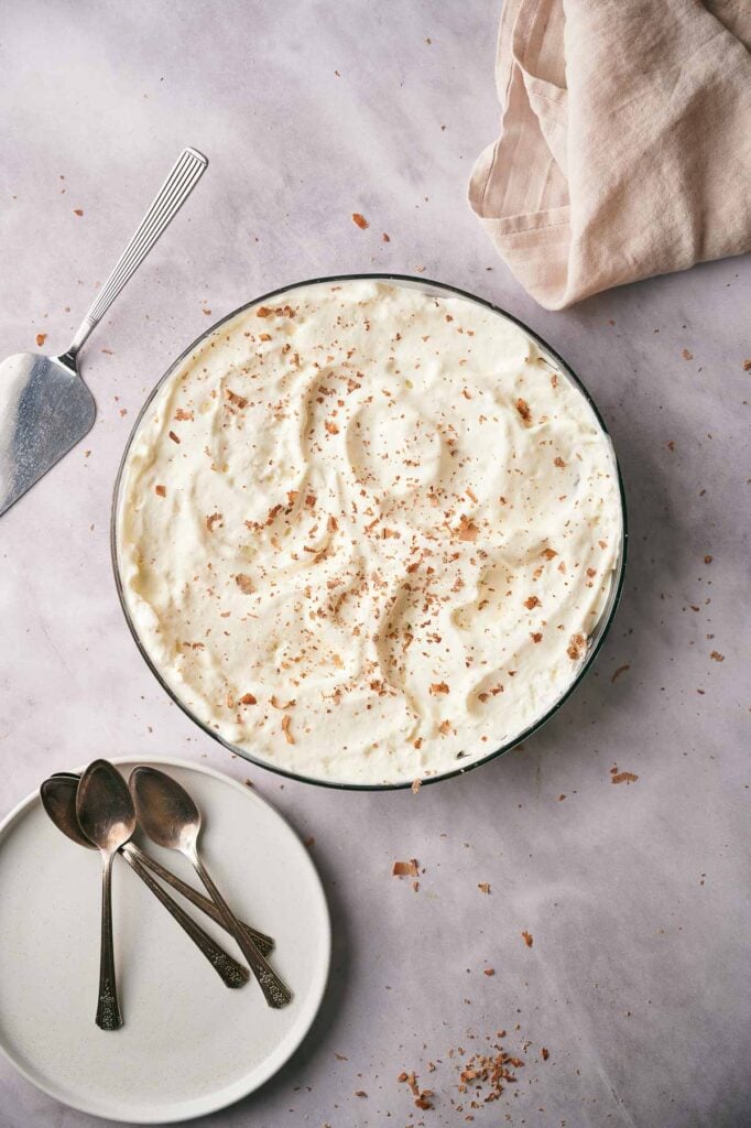 A bowl of creamy Mississippi Mud Pie dessert topped with grated chocolate sits on a light surface beside a pie server, three spoons on a plate, and a beige napkin.