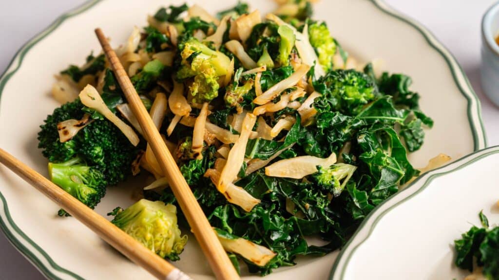 A plate of saut&eacute;ed kale, broccoli, and onions with a pair of wooden chopsticks resting on the rim.