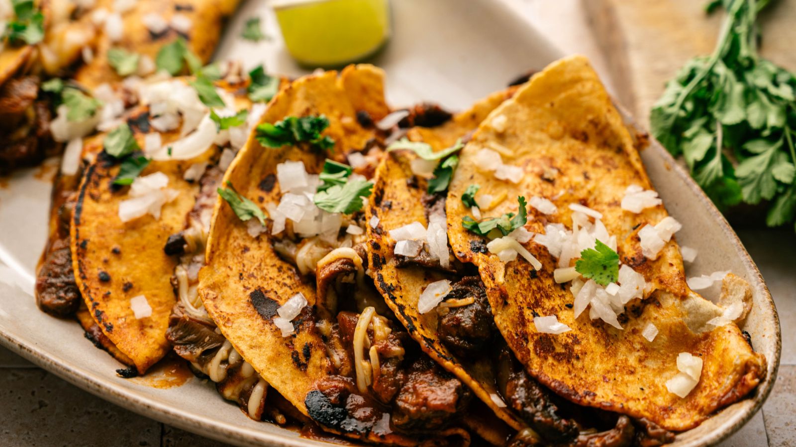 A close-up of three birria tacos on a plate, topped with chopped onions and cilantro, with a lime wedge and fresh herbs in the background.