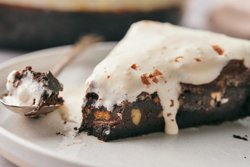 A decadent slice of Mississippi Mud Pie brownie with nuts, topped with melting vanilla ice cream on a plate, served with a spoon beside it.