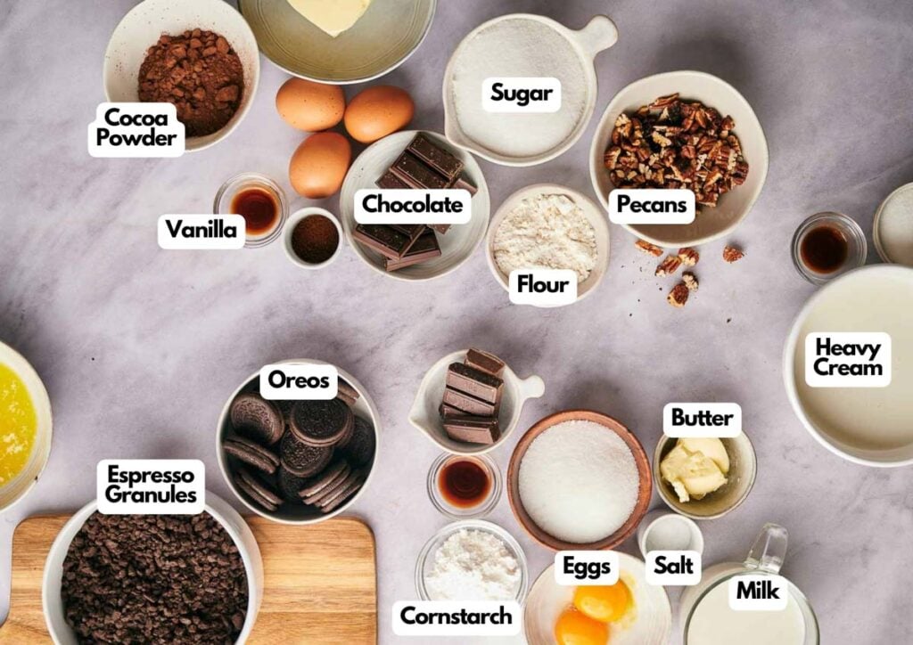 An overhead view of various baking ingredients in bowls—perfect for making a decadent Mississippi Mud Pie—including labeled cocoa powder, sugar, pecans, vanilla, chocolate, flour, heavy cream, Oreos, espresso granules, butter, eggs, cornstarch, salt, and milk.