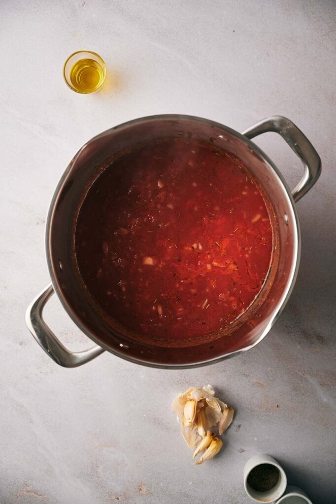 A large pot of pizza sauce sits on a countertop, with a small glass of olive oil, a bulb of garlic, and a pinch of ground pepper nearby.