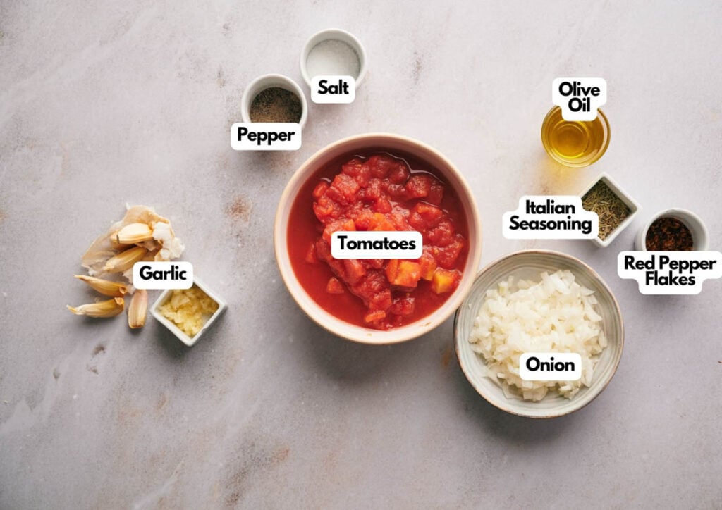 A bowl of chopped tomatoes, diced onion, minced garlic, and small bowls of olive oil, Italian seasoning, red pepper flakes, salt, and pepper are arranged on a white surface&mdash;perfect ingredients for homemade pizza sauce.