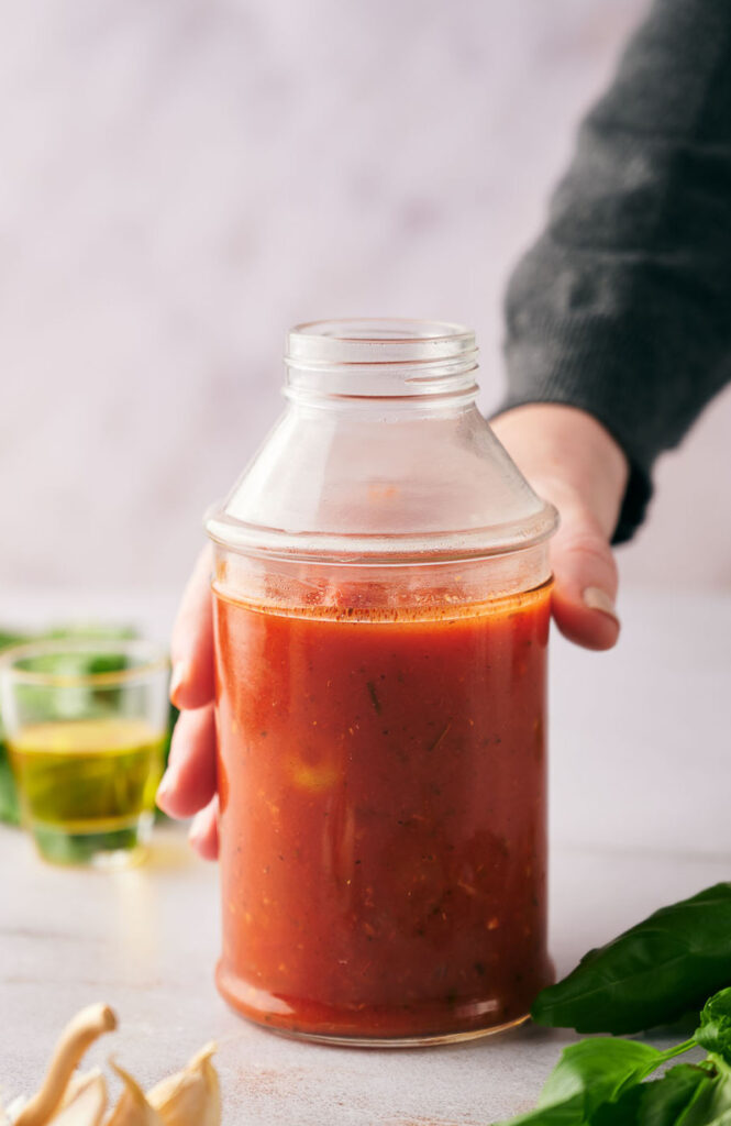 A person holding a glass jar filled with red pizza sauce on a white surface, with greens and a glass of oil in the background.