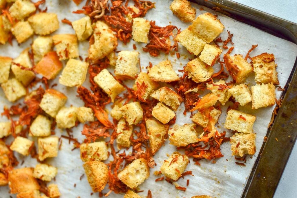 Baking sheet with evenly spread cheese croutons and shredded vegetables on parchment paper.
