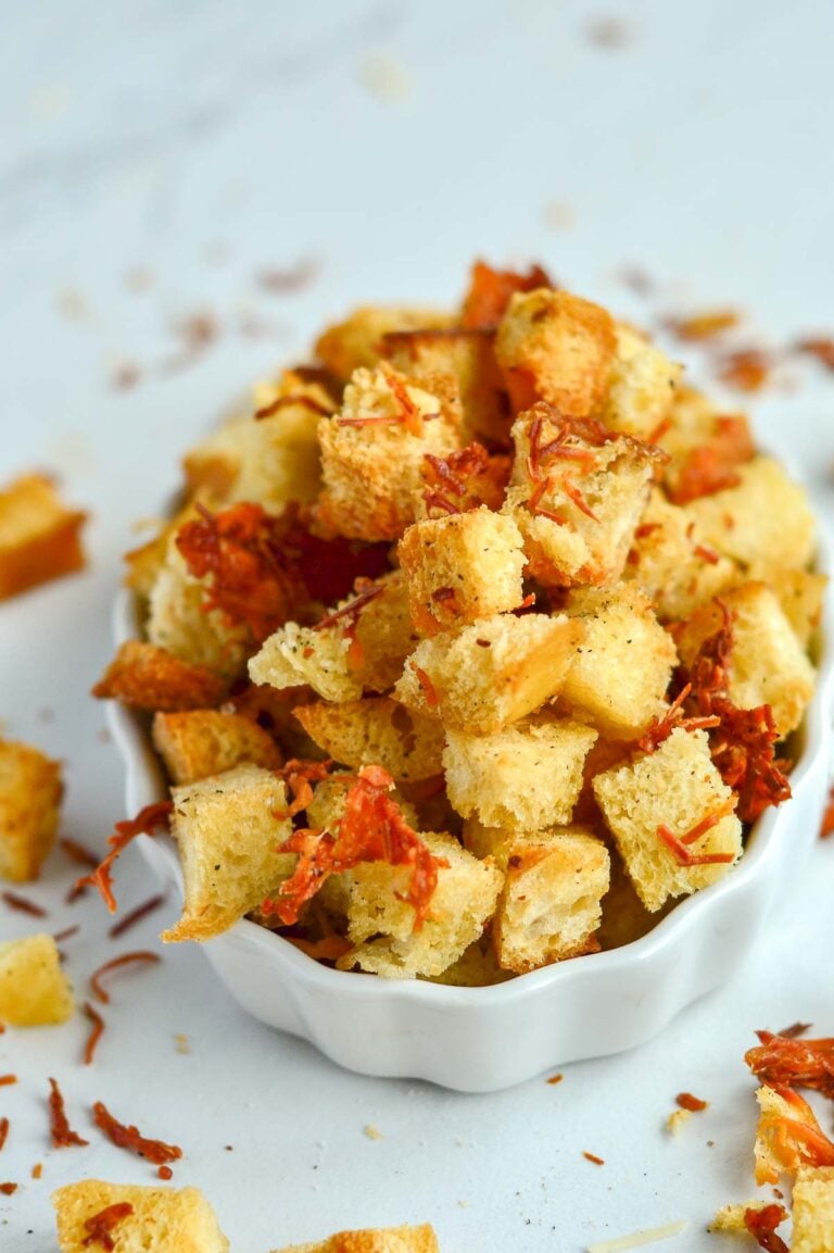 A small white bowl filled with cheese croutons and shredded dried tomato pieces, with some croutons scattered on a white surface.