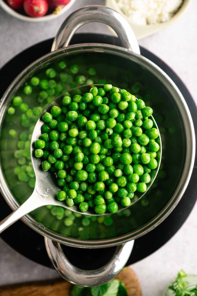 A slotted spoon lifts bright green cooked peas from a pot of water, ready to be chilled.