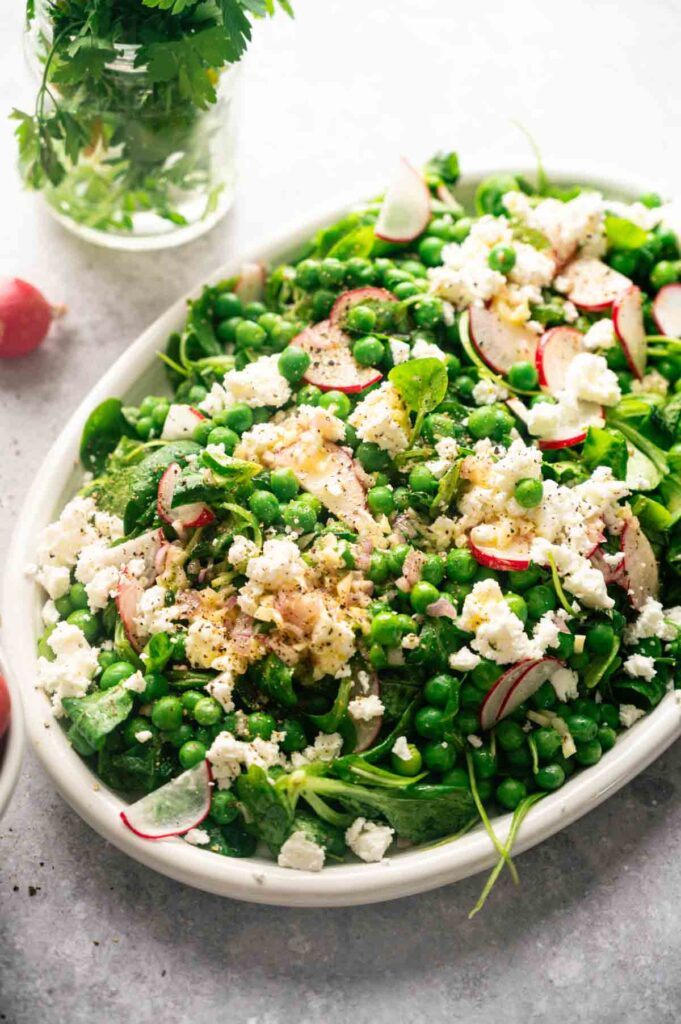 A platter of pea salad is topped with crumbled feta, sliced radishes, and fresh arugula, dressed with lemon maple vinaigrette.