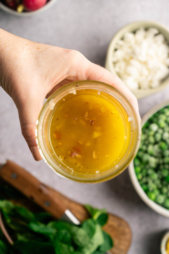 A hand holds a jar of lemon maple dressing with minced shallot and garlic, ready to be shaken together.