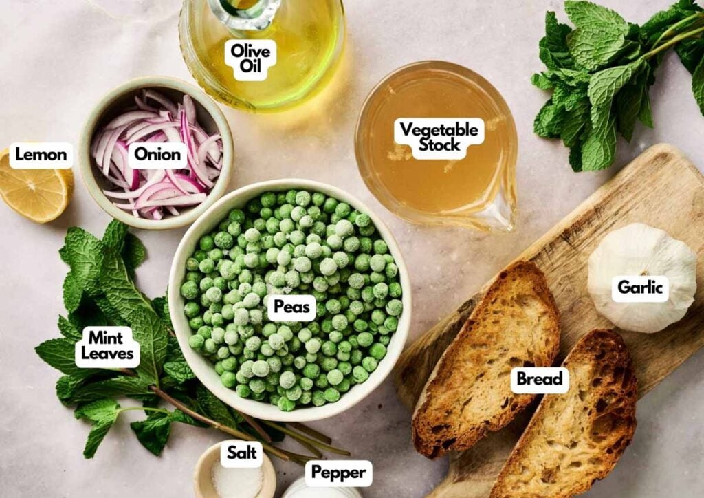 Ingredients for Air Fryer Pea and Mint Soup laid out on a table, including peas, bread, garlic, onion, olive oil, vegetable stock, mint leaves, lemon, salt, and pepper.