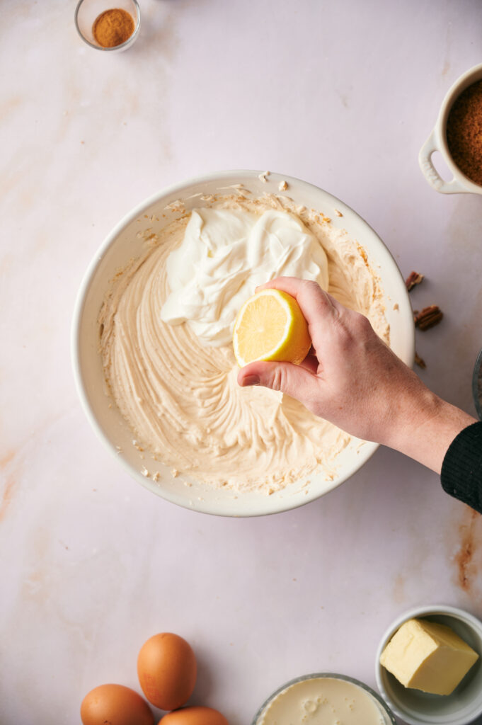 A hand squeezes a lemon half into a bowl of creamy mixture, surrounded by eggs, butter, and other baking ingredients on a light surface—a perfect scene for preparing pecan pie cheesecake.