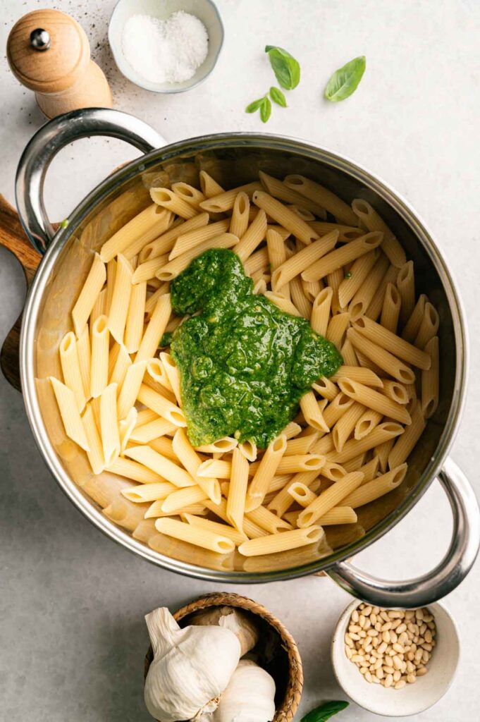 A pot of Pesto Pasta features cooked penne topped with green pesto sauce, surrounded by fresh basil, garlic, pine nuts, salt, and a pepper grinder.
