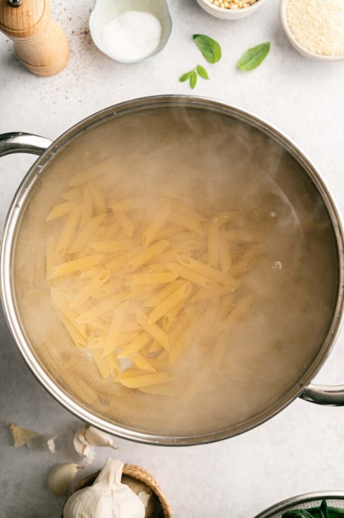 A pot of pasta boiling in water on a stove, surrounded by garlic, salt, and other cooking ingredients on a kitchen counter—perfect preparation for a delicious Pasta al pesto.