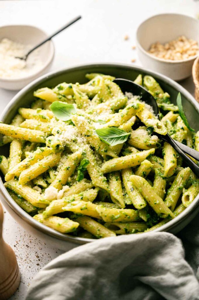 A bowl of Pesto Pasta coated in green pesto sauce, garnished with grated cheese and basil leaves, with a serving spoon and bowls of pine nuts and extra grated cheese nearby.