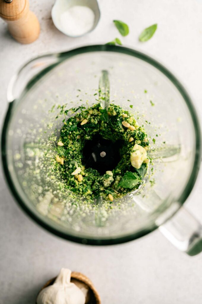Top view of a blender with partially blended pesto pasta ingredients, including basil leaves, garlic, pine nuts, and grated cheese.