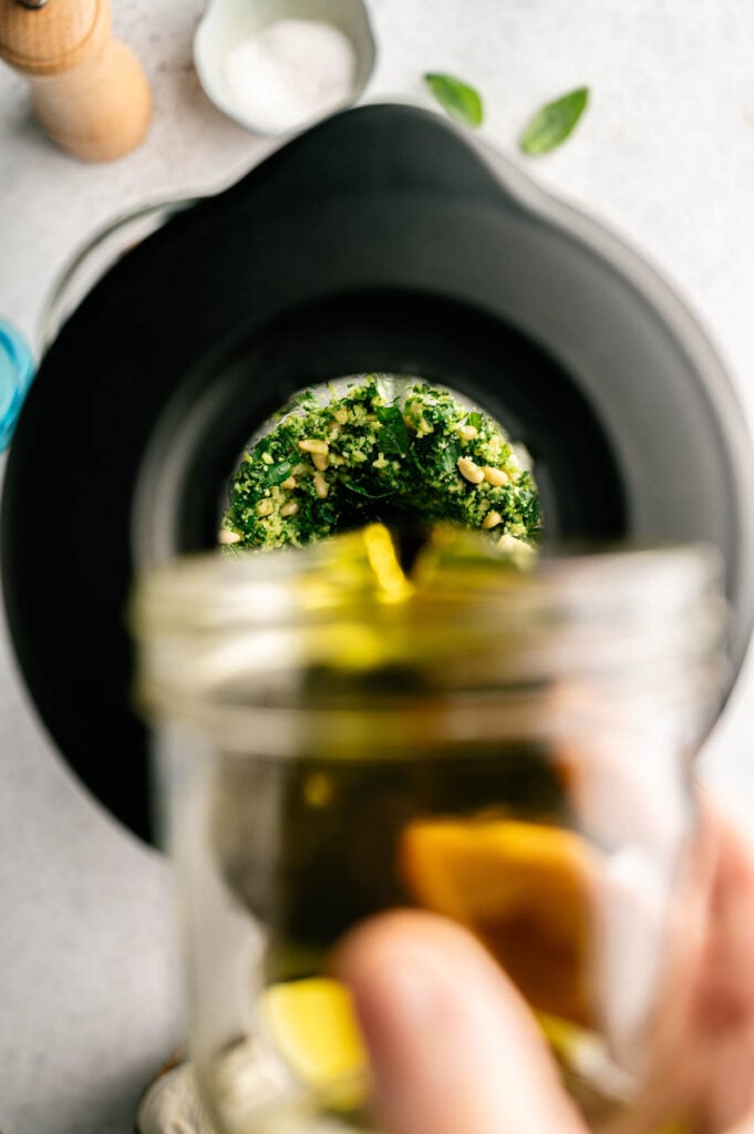 A hand pours olive oil from a jar into a food processor containing chopped greens and nuts, beginning the process of making a fresh Pasta al pesto sauce.
