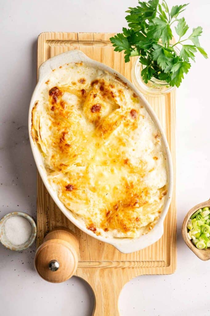 A baked dish of Potatoes Au Gratin sits on a wooden board, surrounded by parsley, green onions, a pepper grinder, and a small bowl of salt.