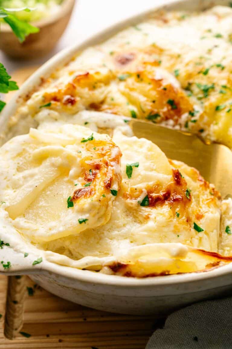 Close-up of creamy Potatoes Au Gratin baked with cheese and garnished with chopped parsley, served with a golden spatula in a white casserole dish.