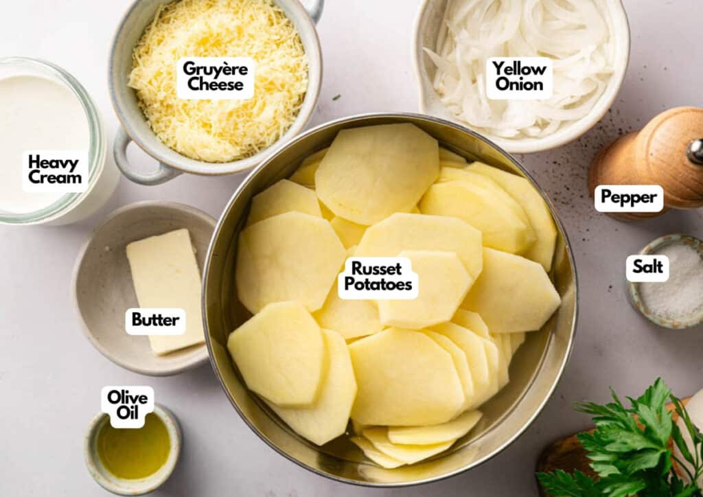 A top view of labeled ingredients for Potatoes Au Gratin, including russet potatoes, Gruy&egrave;re cheese, yellow onion, heavy cream, butter, olive oil, pepper, salt, and parsley.