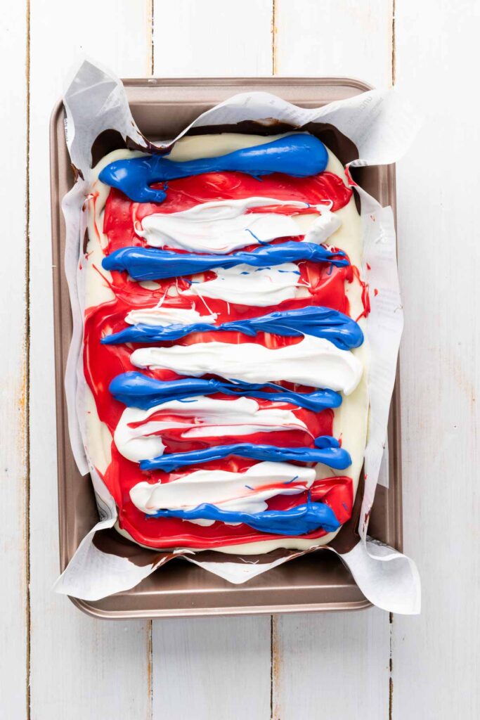 A pan of layered Red White and Blue Chocolate Bark dessert with vibrant red, white, and blue melted chocolate drizzled in stripes, lined with parchment paper on a white wooden surface.