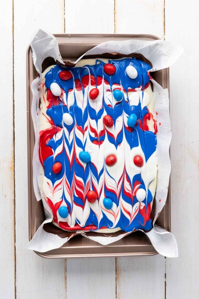 A baking pan lined with parchment paper holds a Red White and Blue Chocolate Bark dessert, topped with swirled icing and matching candy pieces.