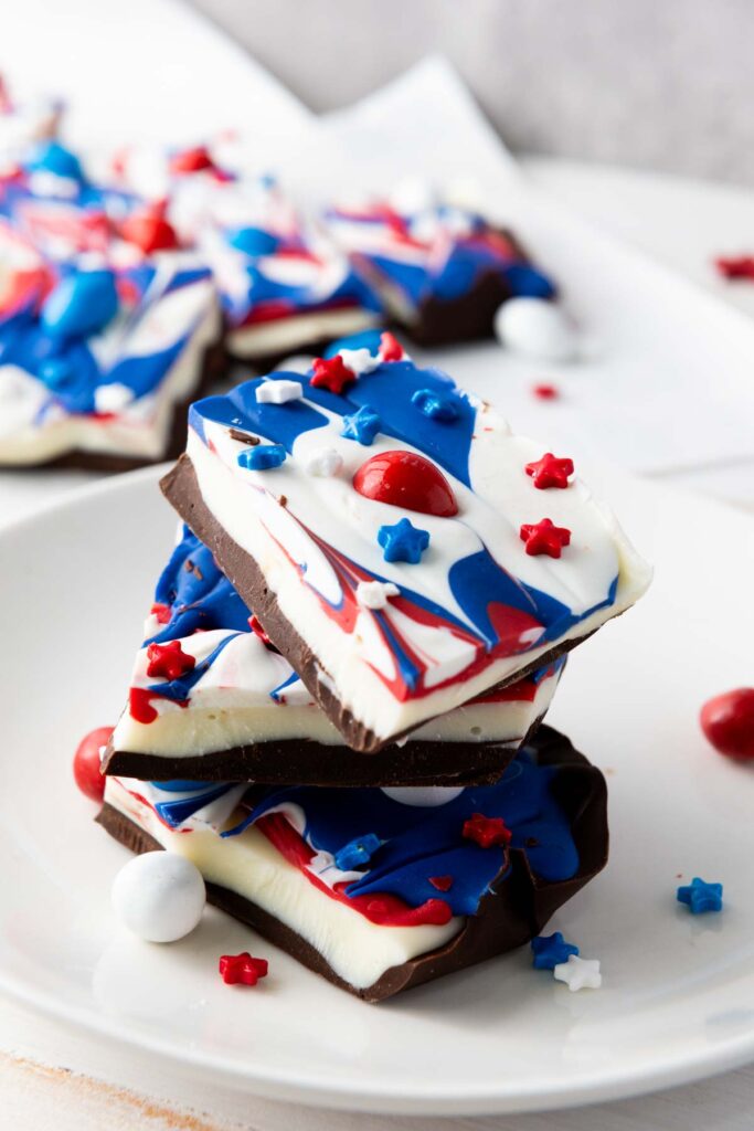 Three pieces of Red White and Blue Chocolate Bark topped with vibrant candy melts and star sprinkles, stacked on a white plate, make a festive treat for any patriotic celebration.