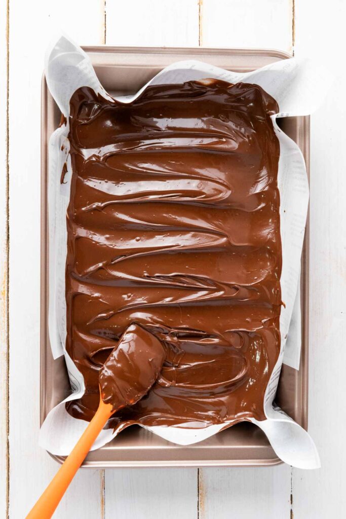 A baking pan lined with parchment paper holds a smooth layer of melted chocolate being spread with an orange spatula, the perfect base for creating festive Red White and Blue Chocolate Bark.
