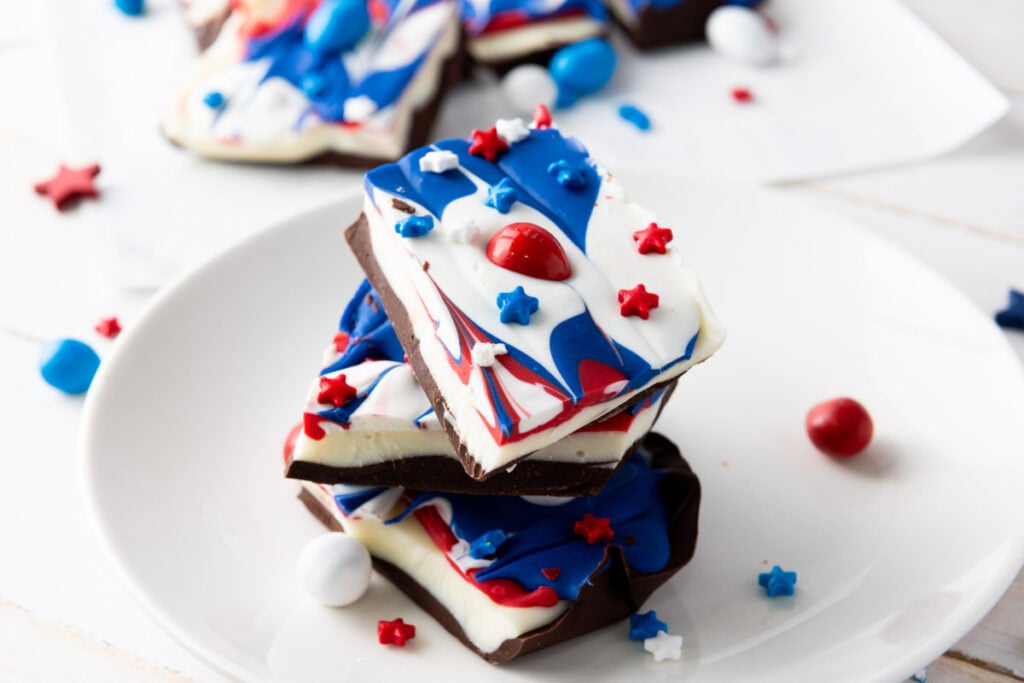 Stack of Red White and Blue Chocolate Bark pieces with swirled patriotic toppings, decorated with star-shaped sprinkles and candy-coated chocolates on a white plate.