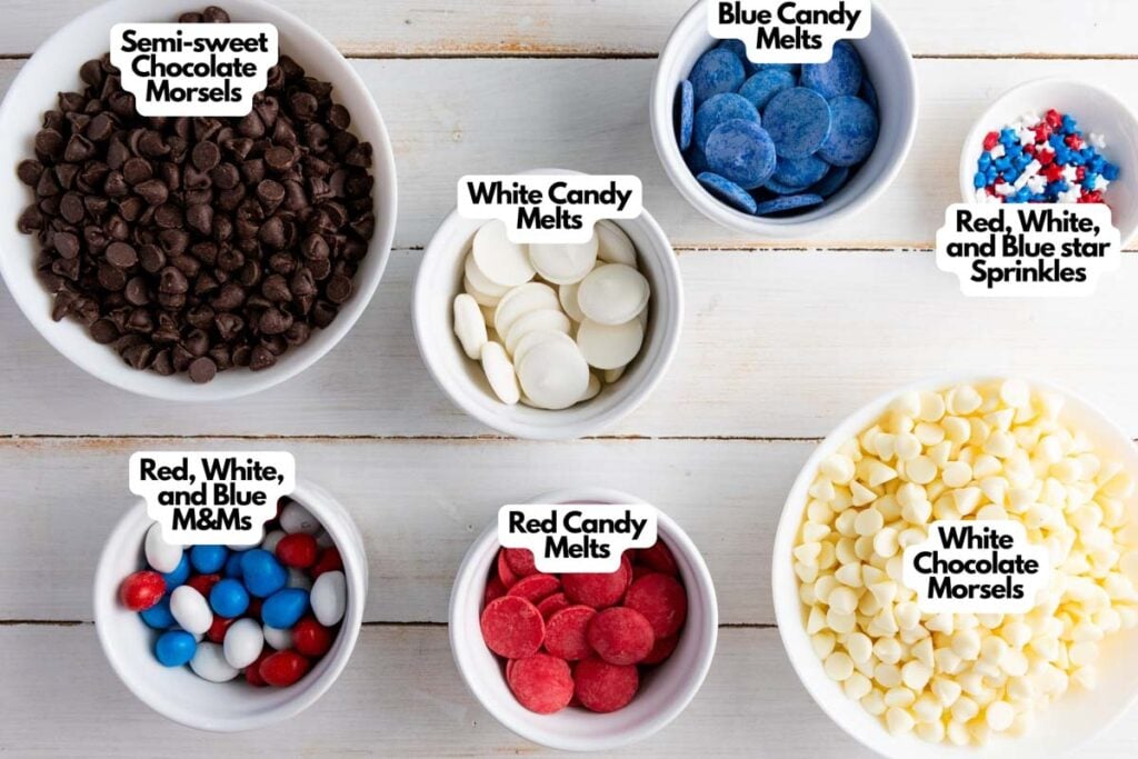 Six bowls hold the ingredients for Red White and Blue Chocolate Bark: semi-sweet chocolate morsels, white and blue candy melts, star sprinkles, M&Ms, red candy melts, and white chocolate morsels.
