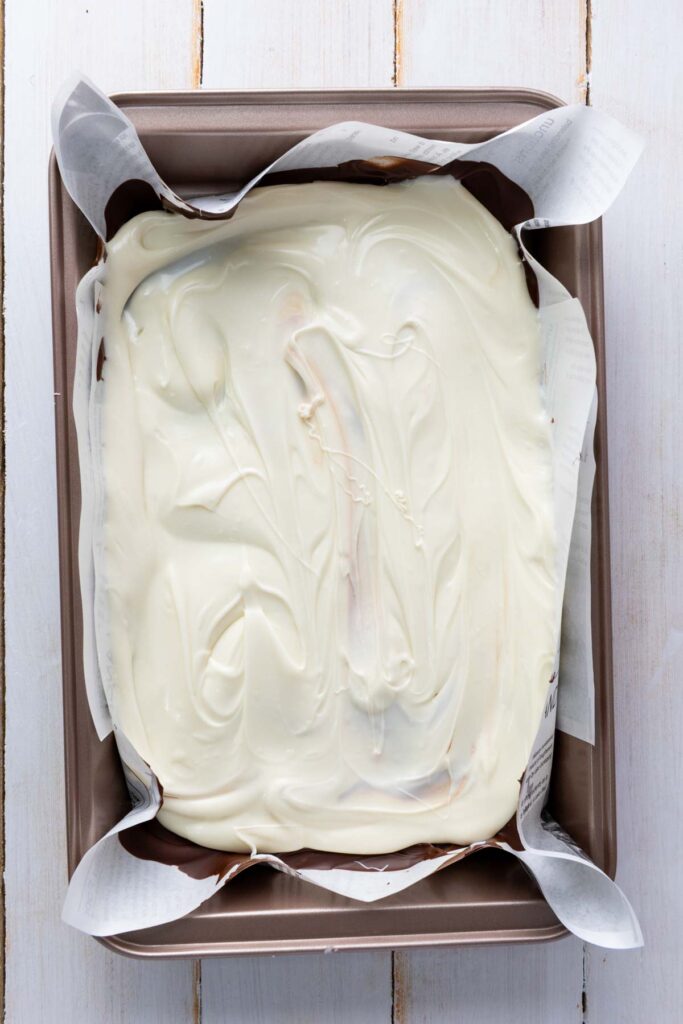A smooth layer of white chocolate covers a dark chocolate base in a lined pan, the swirled surface still soft before it sets.